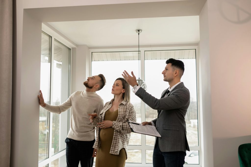 A real estate agent in a suit shows a modern, bright apartment to a young couple, one of whom is pregnant. The agent gestures toward the large windows while the couple looks up, examining the property.